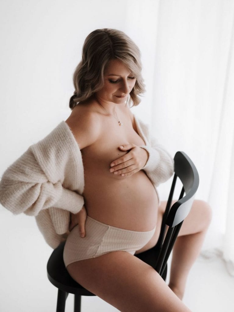 Schwangere trägt nur Slip und Strickjacke und sitzt auf einem Stuhl