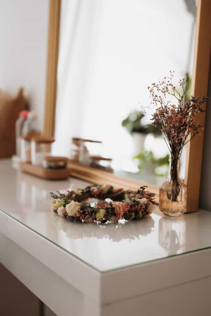 Schöner Blumenkranz und Blumen in einer kleinen Vase stehen vor einem Spiegel auf weißem Tischchen.