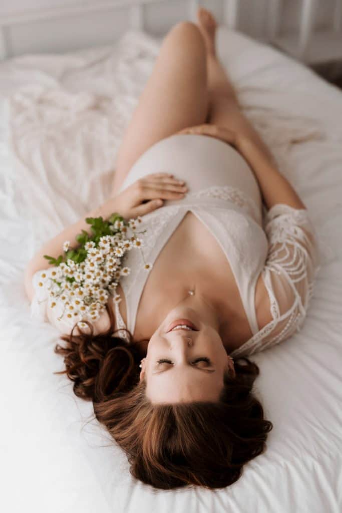 Schwangere Frau liegt mit Kamilleblüten im Arm auf dem Bett, sie trägt einen weißen Body und streichelt mit ihren Händen ihren Babybauch.