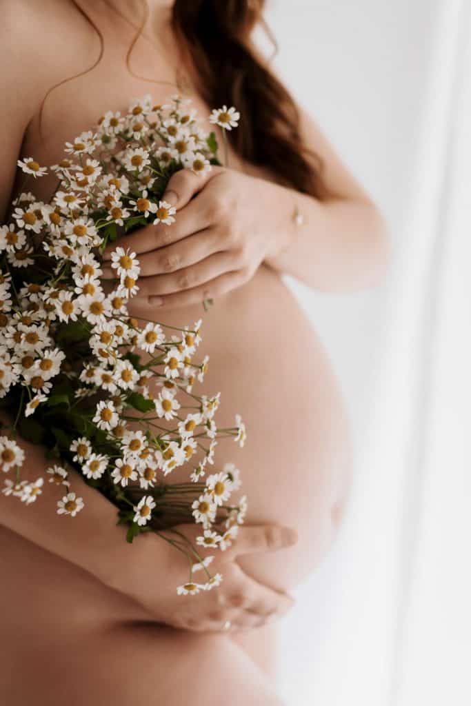 Schwangere Frau steht unbekleidet, seitlich zur Kamera, bedeckt ihre Brust mit der Hand und Kamilleblüten und hält mit der anderen Hand ihren Babybauch.