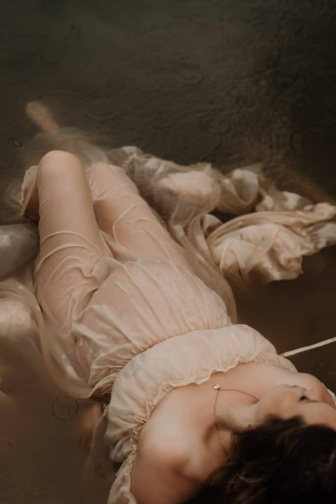Eine schwangere Frau liegt in einem nassen, transparenten Kleid im Wasser, das ihren zauberhaften Babybauch zart umhüllt.