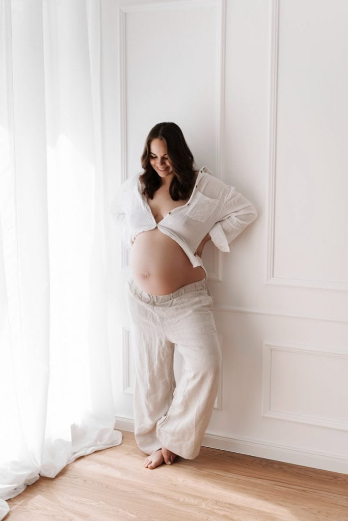 Schwangere Frau in beiger Leinenhose und weißem Hemd, bei welchem nur der oberste Knopf geschlossen ist, sodass der Babybauch zu sehen ist, steht mit den Händen in den Hüften und blickt lächelnd auf ihren Babybauch hinab.