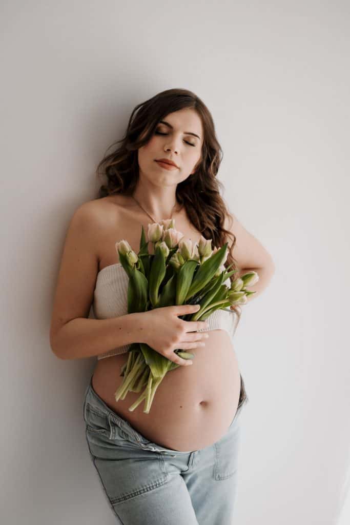 Schwangere Frau mit Tulpen im Arm lehnt an der Wand, hat die Augen geschlossen und zeigt ihren runden Babybauch.