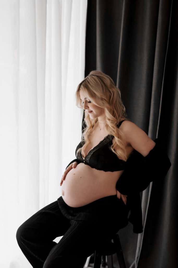 Schwangere Frau mit langen, lockigen blonden Haaren und schwarzen Dessous sitzt seitlich auf einem Hocker und streichelt verträumt ihren Babybauch.
