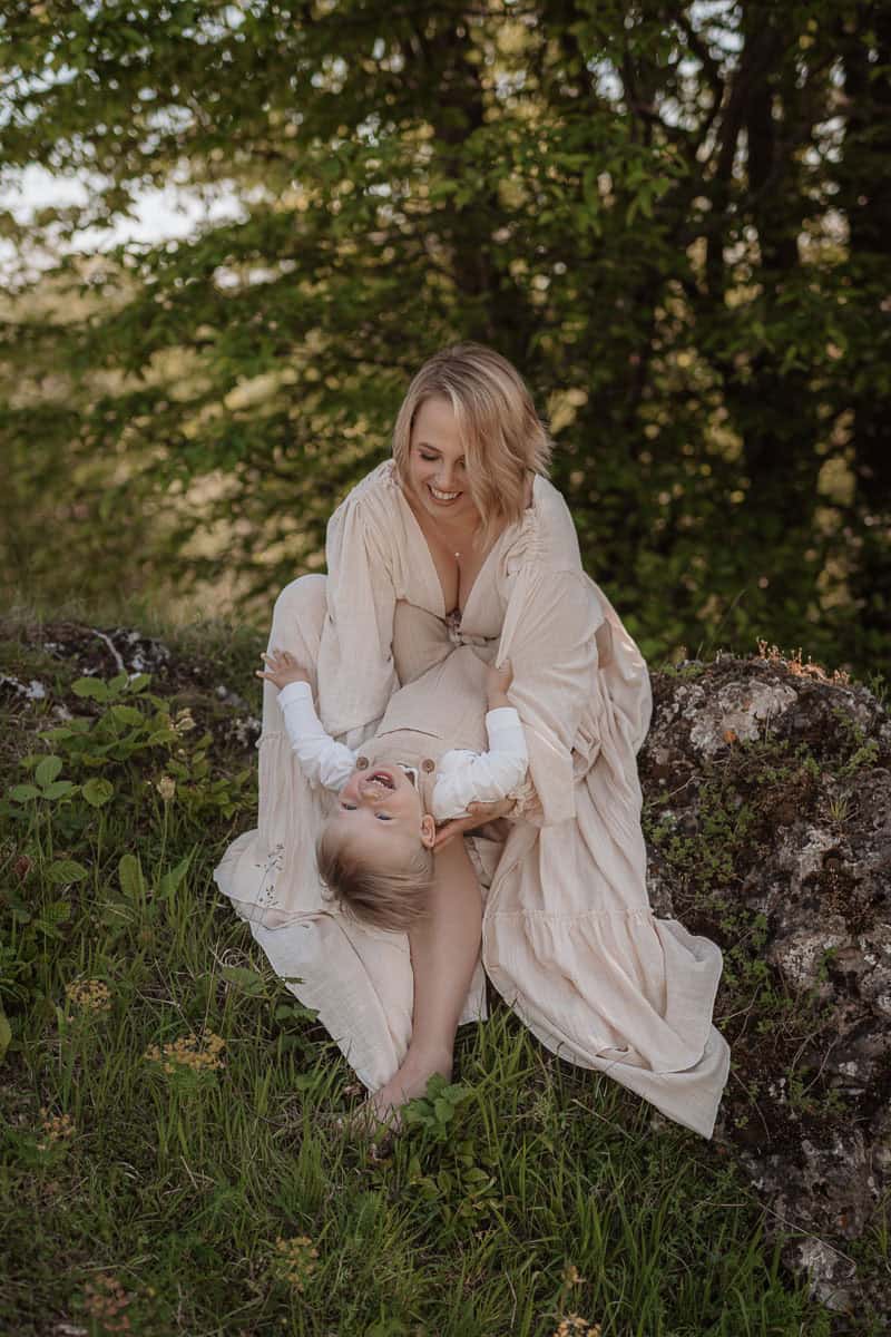 Eine blonde Frau sitzt lächelnd auf einer Wiese inmitten von grünem Laub und spielt liebevoll mit einem kleinen Kind. Die Frau trägt ein langes, cremefarbenes Kleid, das sich sanft um sie legt. Das Kind, in einem weißen Oberteil, lacht ausgelassen und liegt auf ihrem Schoß, während die beiden einen fröhlichen Moment teilen. Die natürliche Umgebung und die innige Verbindung strahlen Wärme und Lebensfreude aus.