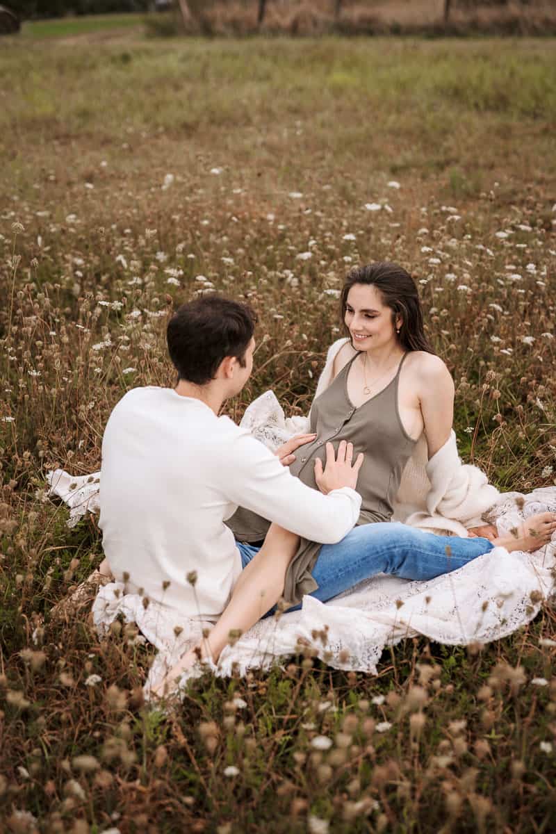 Ein verliebtes Paar sitzt inmitten einer blühenden Wiese auf einer Decke. Der Mann trägt einen weißen Pullover und Jeans, während die Frau ein khakifarbenes Kleid trägt und eine leichte Strickjacke um die Schultern gelegt hat. Beide lächeln sich an, während er liebevoll seine Hände auf ihren Babybauch legt. Die natürliche Umgebung vermittelt eine warme und intime Atmosphäre.