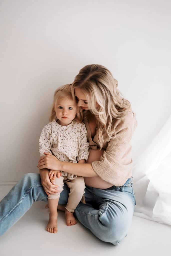 Eine schwangere Frau sitzt entspannt auf dem Boden in einer hellen, minimalistischen Umgebung. Sie trägt eine lockere Bluse und Jeans und hält liebevoll ein kleines Mädchen auf ihrem Schoß. Das Kind trägt ein gemustertes Outfit und schaut mit großen Augen in die Kamera, während die Mutter sanft den Kopf des Mädchens berührt und dabei lächelt. Eine warme und intime Atmosphäre wird durch das weiche Licht und die natürliche Umgebung geschaffen.