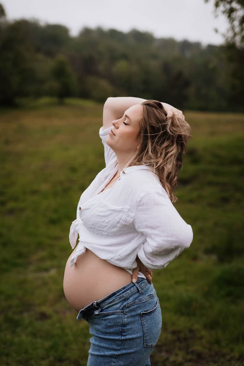 Eine schwangere Frau steht in einer natürlichen, grünen Landschaft. Sie trägt eine aufgeknöpfte weiße Bluse und eine Jeans, wobei ihr Babybauch liebevoll betont wird. Mit geschlossenen Augen lehnt sie ihren Kopf leicht zurück und fährt mit einer Hand durch ihr Haar, während die andere Hand ihren unteren Rücken stützt. Die Szene wirkt ruhig und kraftvoll, untermalt von der sanften, bewölkten Atmosphäre der Umgebung.