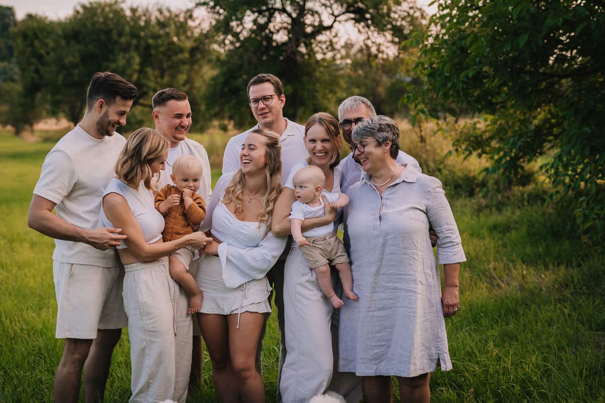 Eine große, fröhliche Familie steht in einem grünen Feld unter Bäumen und genießt einen gemeinsamen Moment. Alle lachen und blicken liebevoll zueinander. Zwei Babys sind im Mittelpunkt des Bildes, getragen von ihren Eltern, während Großeltern und weitere Familienmitglieder die Szene umrahmen. Die Atmosphäre ist herzlich und natürlich, mit sanftem Abendlicht im Hintergrund.