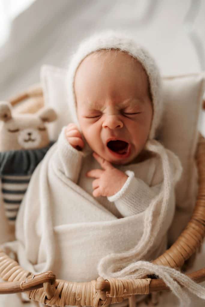 Ein neugeborenes Baby liegt in einem kleinen geflochtenen Korb, eingewickelt in eine weiche cremefarbene Decke. Es trägt eine gestrickte, weiße Mütze und gähnt herzhaft mit geschlossenen Augen. Im Hintergrund liegt ein Plüschspielzeug in Form eines Bären, das die Szene liebevoll ergänzt. Die Beleuchtung ist sanft und unterstreicht die Ruhe und Zartheit des Moments.