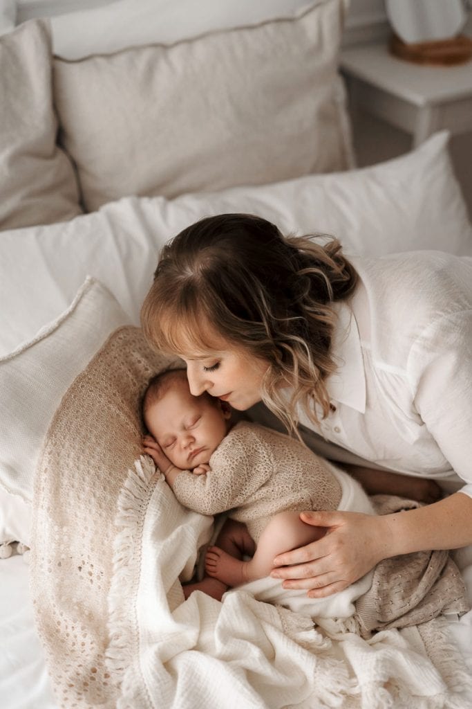 Eine Mutter liegt liebevoll über ihr friedlich schlafendes Baby gebeugt. Das Neugeborene trägt einen gestrickten Body und ist in sanfte Decken gebettet. Die Mutter, in einem weißen Oberteil, strahlt Zärtlichkeit aus, während sie sanft das Baby berührt. Die Szene ist in warmen, neutralen Tönen gehalten und vermittelt Geborgenheit und Nähe.