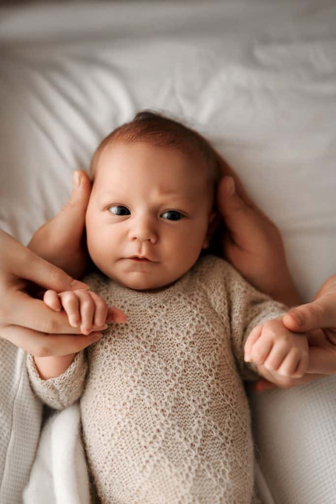 Ein Neugeborenes liegt auf einem weißen Bett, umgeben von warmem, natürlichem Licht. Die kleinen Hände des Babys werden liebevoll von Erwachsenen gehalten, während der Blick des Babys neugierig und aufmerksam in die Kamera gerichtet ist. Das gestrickte Outfit verleiht der Szene eine gemütliche Atmosphäre.