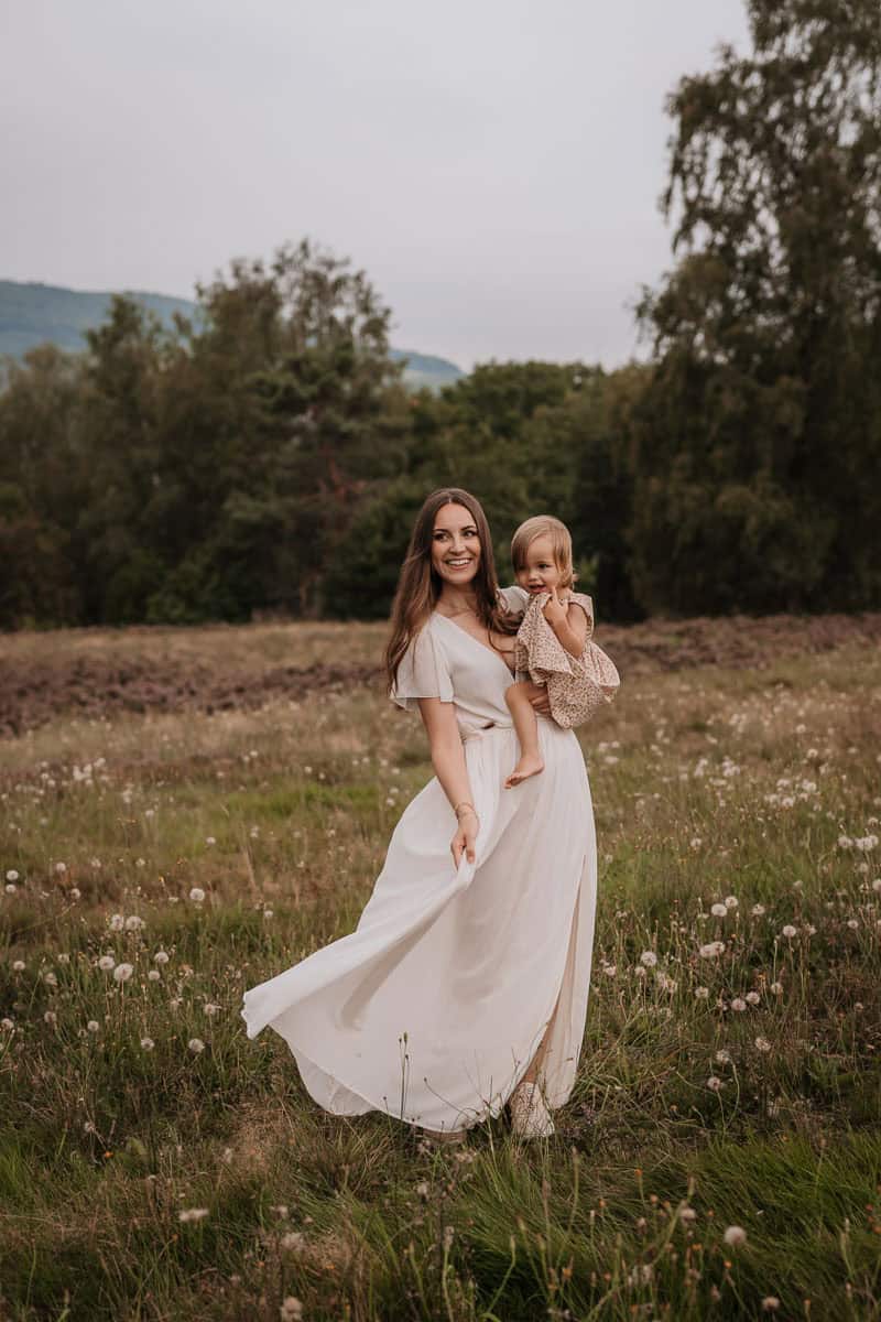 Eine lächelnde Mutter steht auf einer blühenden Wiese und hält liebevoll ihr kleines Kind auf dem Arm. Sie trägt ein fließendes, weißes Kleid, das sanft im Wind weht. Im Hintergrund sind grüne Bäume und Hügel zu sehen, die eine idyllische, natürliche Atmosphäre schaffen.