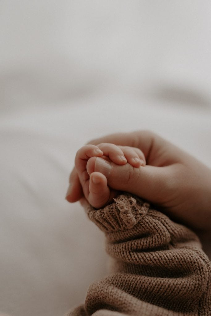 Eine zarte Babyhand greift nach den Fingern einer erwachsenen Hand. Das Baby trägt einen gestrickten, braunen Ärmel, während die Haut der erwachsenen Hand sanft die kleine Hand umschließt.