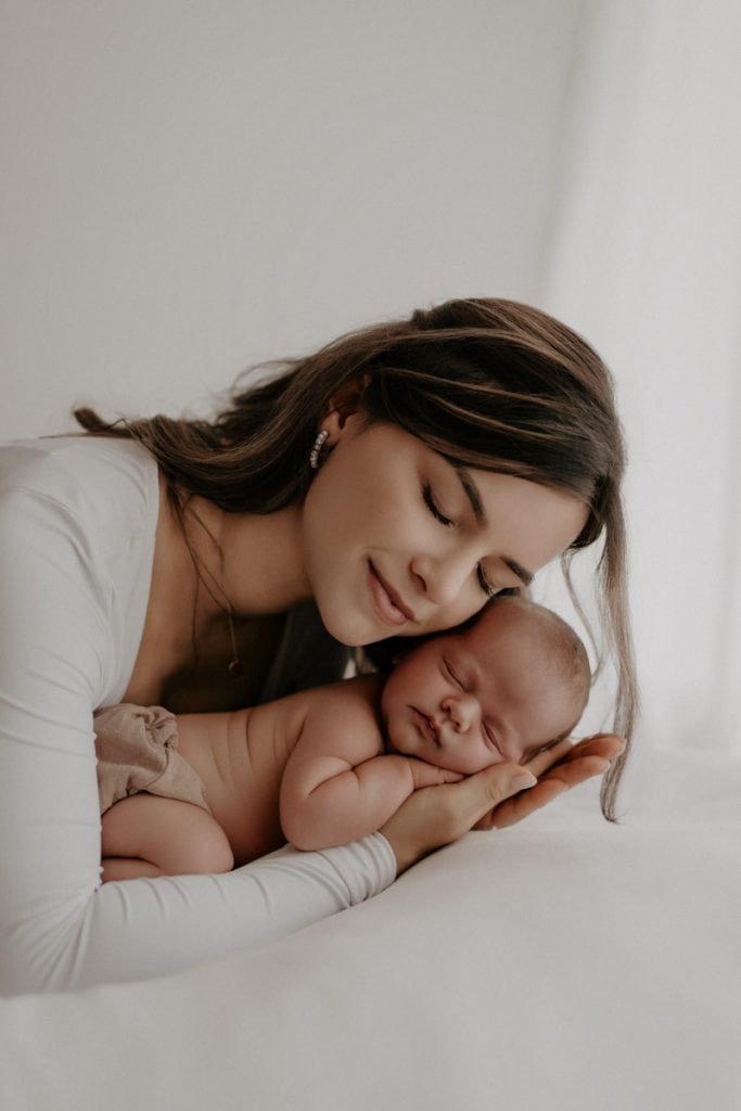 Ein Neugeborenes schläft friedlich auf dem Arm und der Hand seiner Mutter. Sie legt sanft ihren Kopf an das des Babys und hat dabei die Augen geschlossen, wodurch der Moment voller Ruhe und Geborgenheit wirkt.