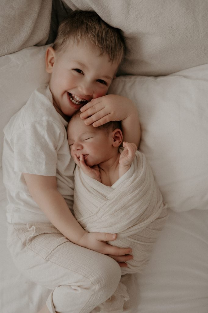 Ein lachender großer Bruder liegt auf einem Bett und hält sein neugeborenes Geschwisterlichen liebevoll im Arm. Das Baby ist in ein weiches, cremefarbenes Tuch gewickelt und schläft friedlich, während sein Bruder es sanft berührt.